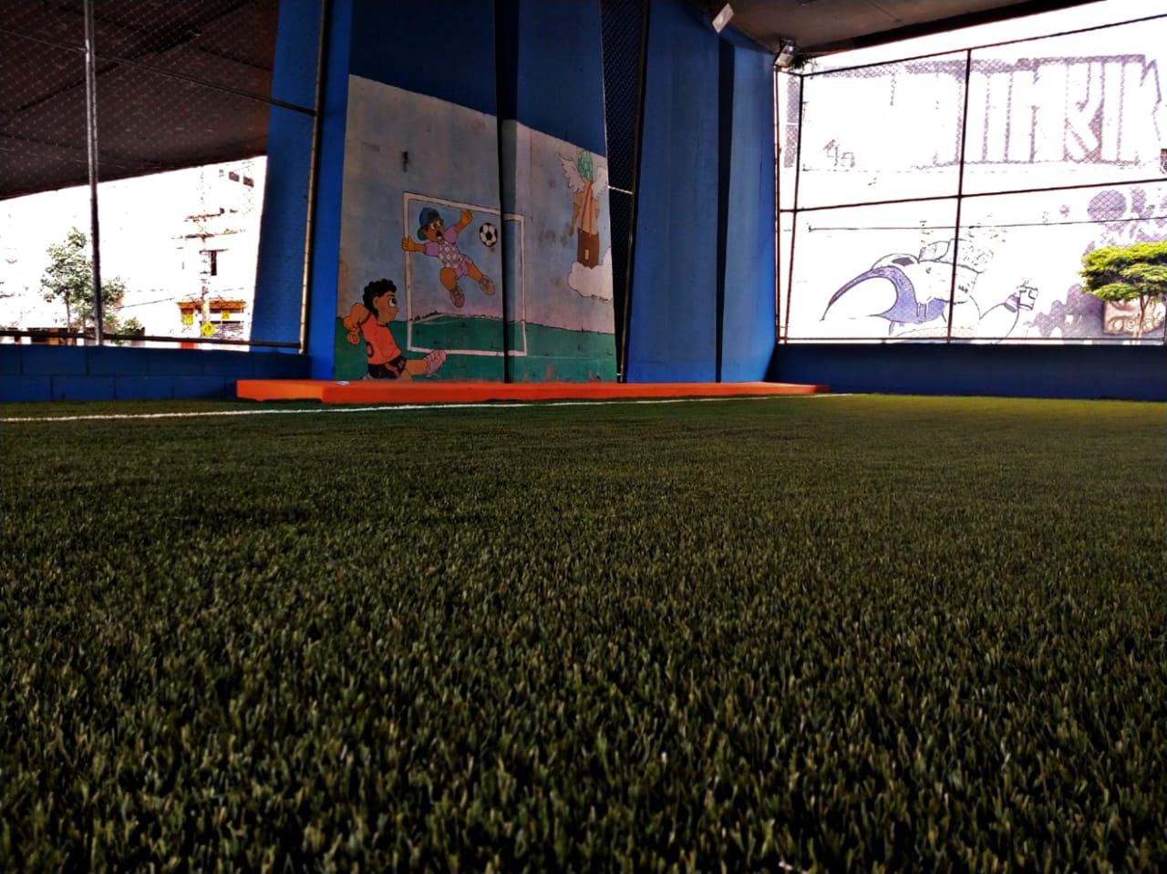 Fotografia colorida mostra gramado da Arena Bela Vista e alambrado é possível visualizar uma pilastra pintada de azul e com o desenho de crianças jogando futebol. 